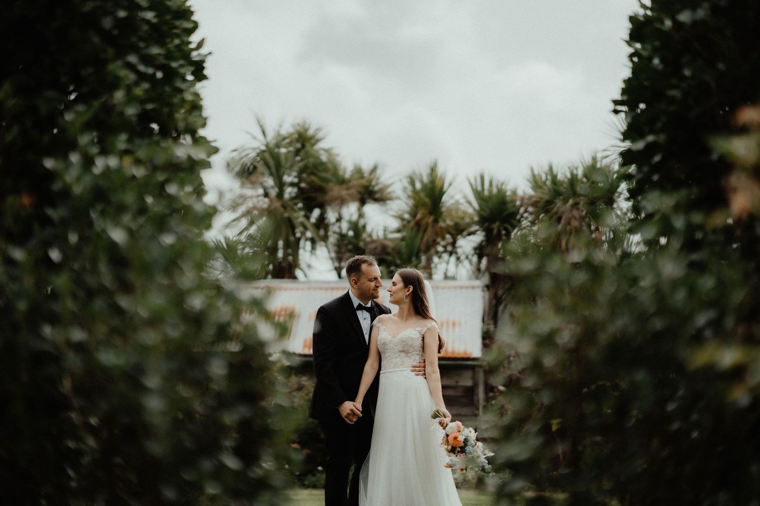 A groom in a black suit embraces his bride in a flowing white gown, holding a bouquet among lush garden greenery and palms.