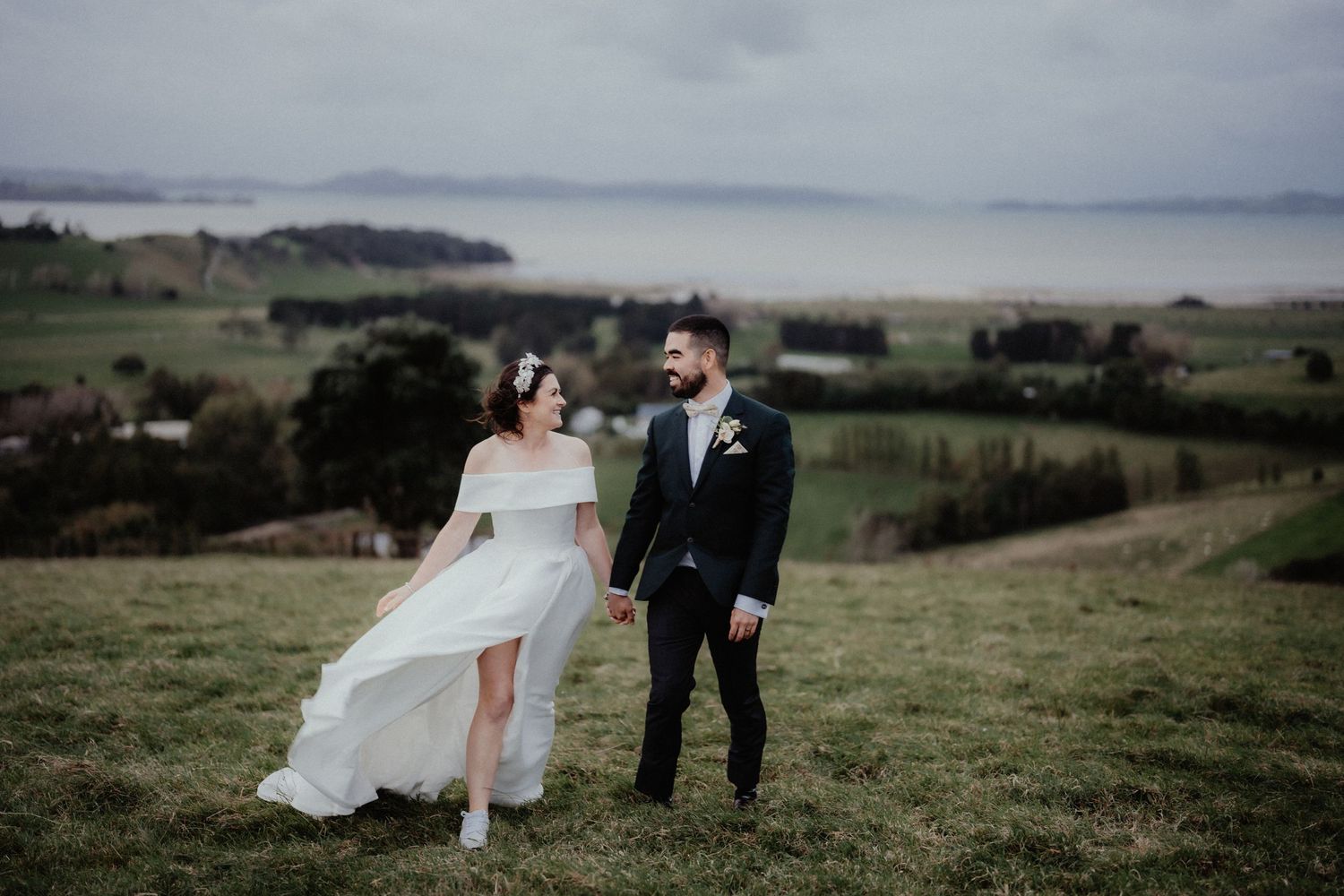 Bride in a flowing white gown and groom in a dark suit walk hand in hand on a grassy hill overlooking the sea.