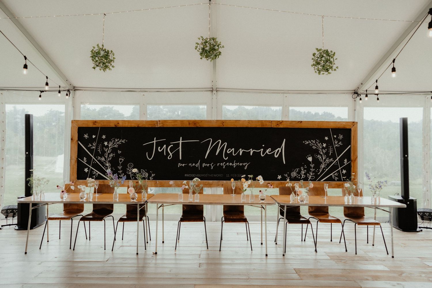 Long reception table set beneath a chalkboard sign reading Just Married inside a bright marquee with hanging greenery.