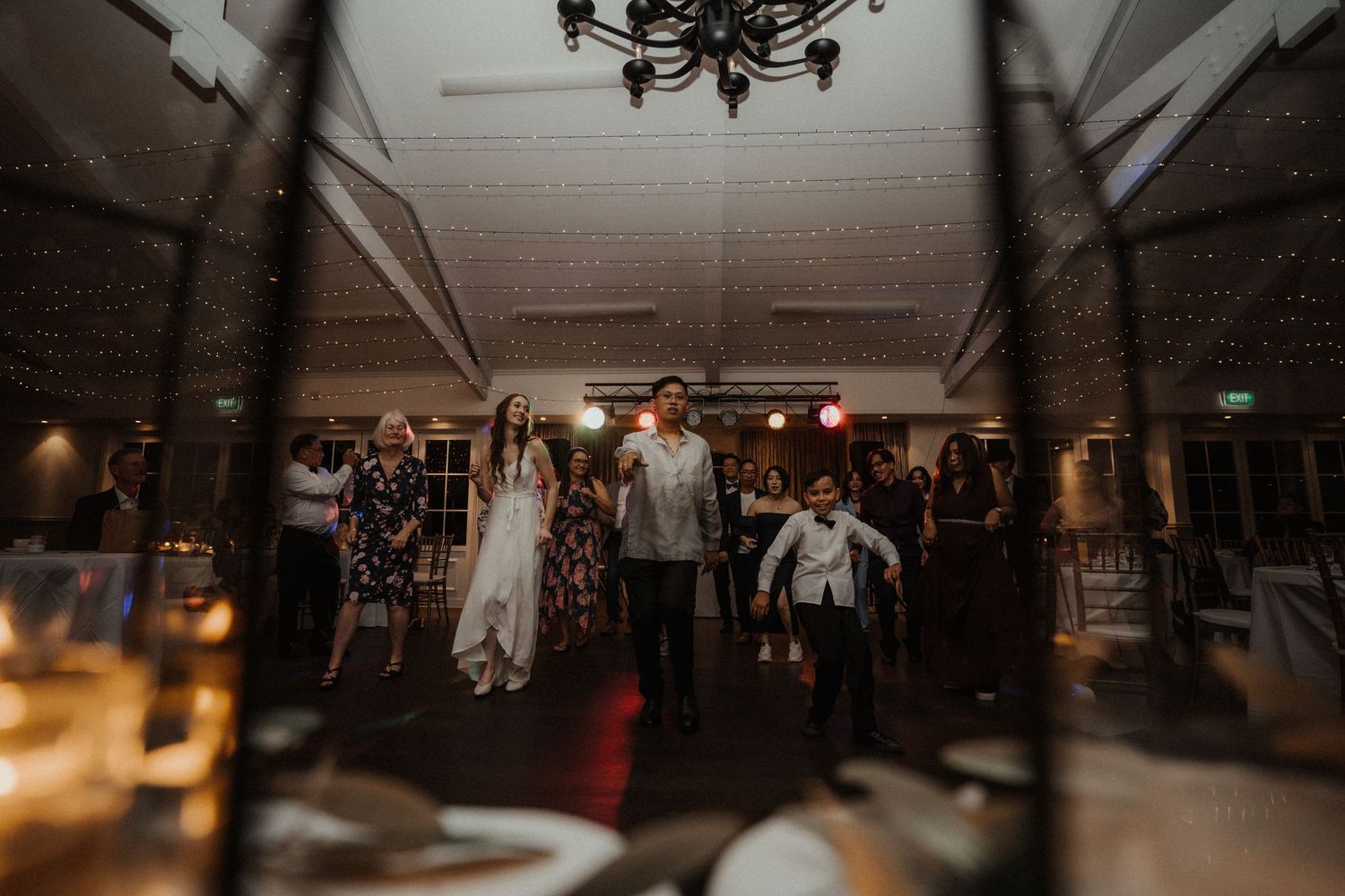 Wedding guests and a bride in a white gown gather on a dimly lit dance floor under a chandelier at an evening reception.