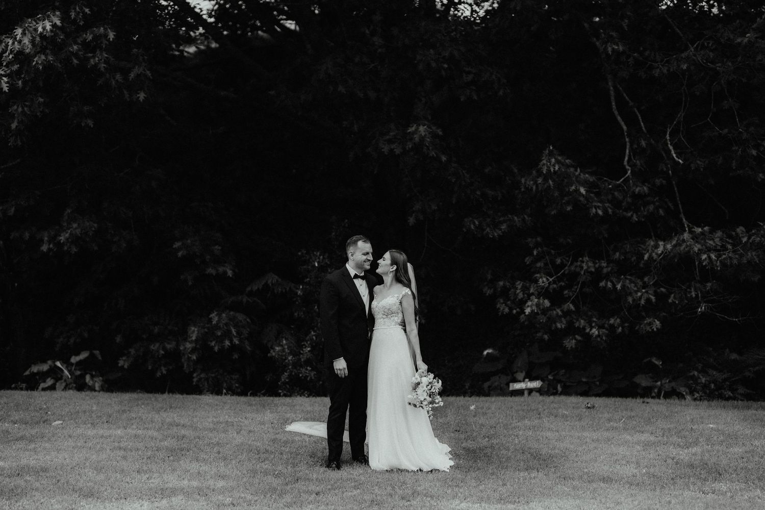 Black and white wedding portrait of a bride and groom embracing on an open lawn in front of dark, dense trees.