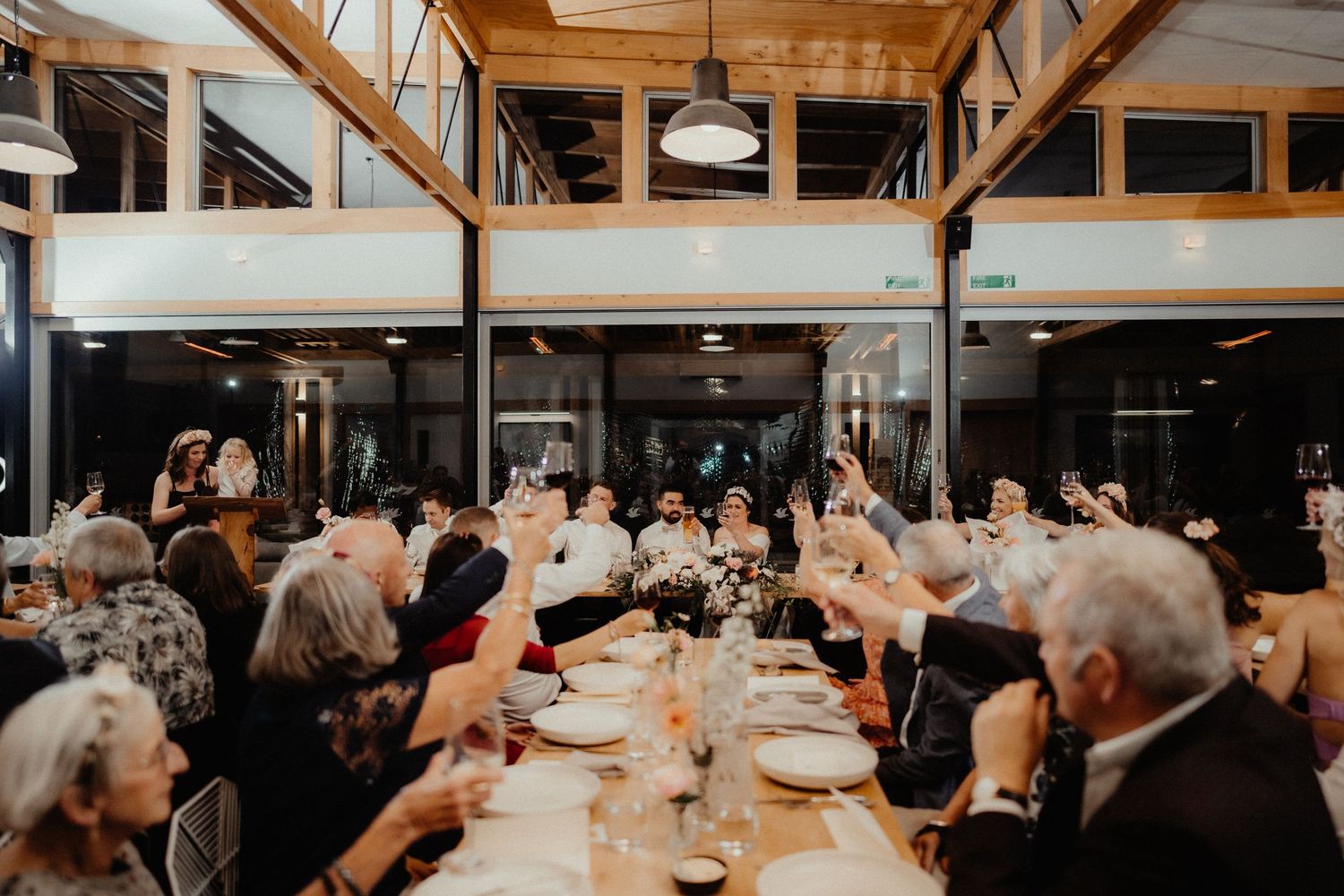 Wedding guests seated at a long banquet table raise their arms and glasses in a cheer inside a rustic timber-beamed venue.