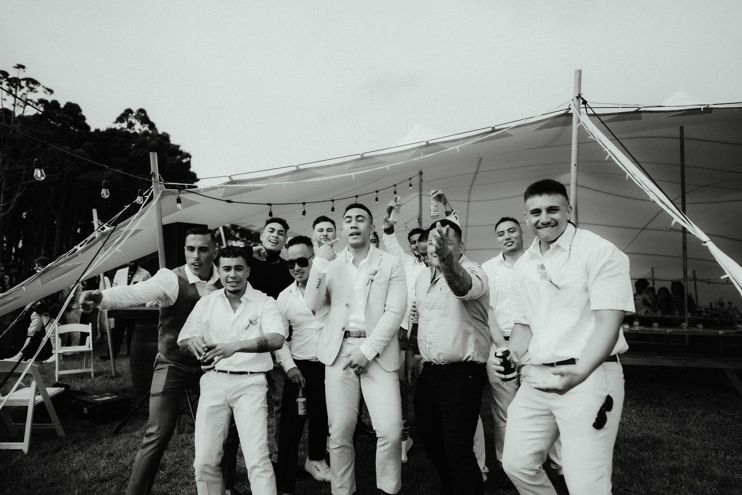 Black and white photo of cheerful groomsmen laughing and celebrating together under a marquee tent at a wedding reception.