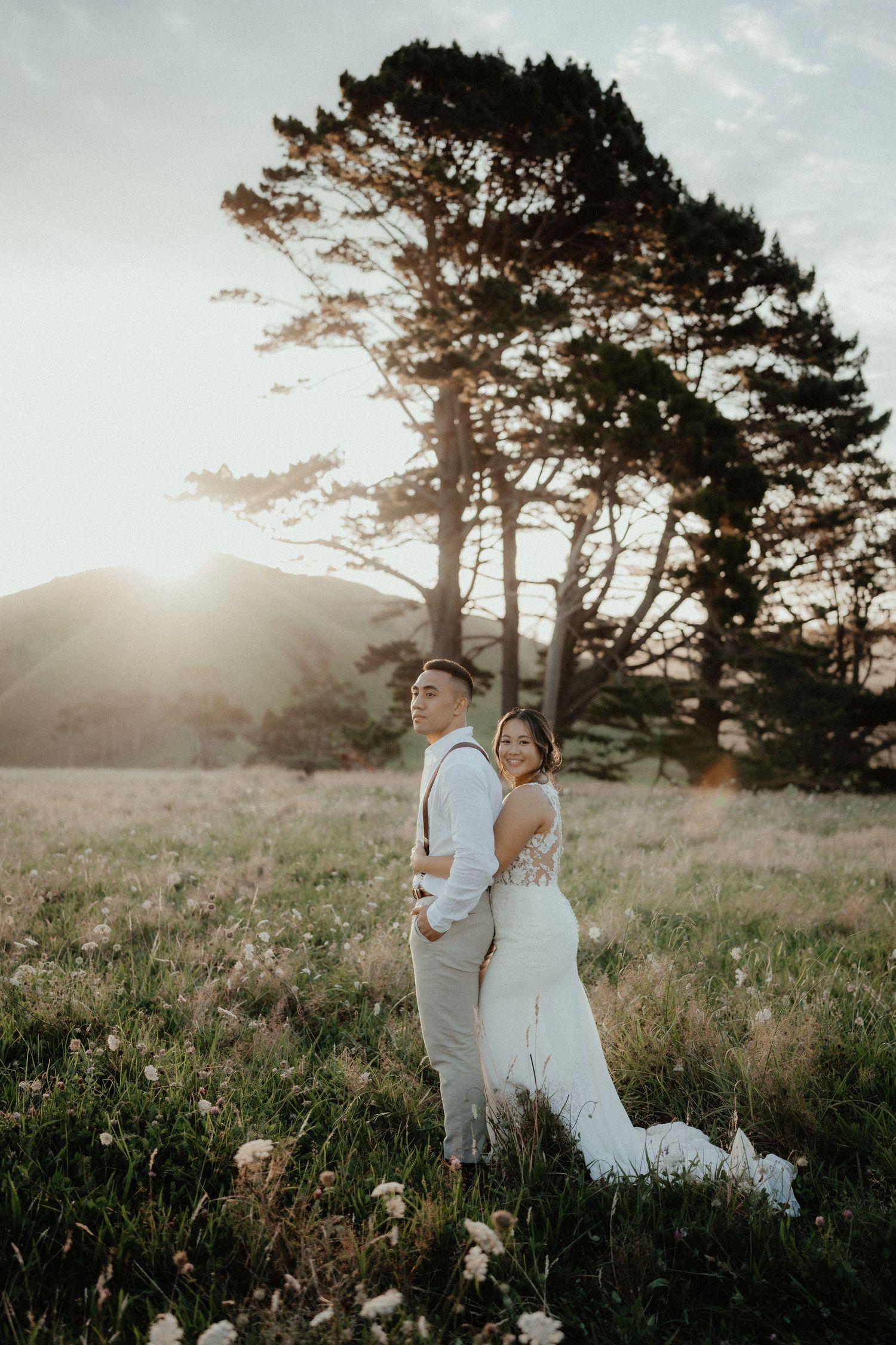 A bride and groom stand in a wildflower meadow at sunset with tall pine trees and rolling hills behind them.