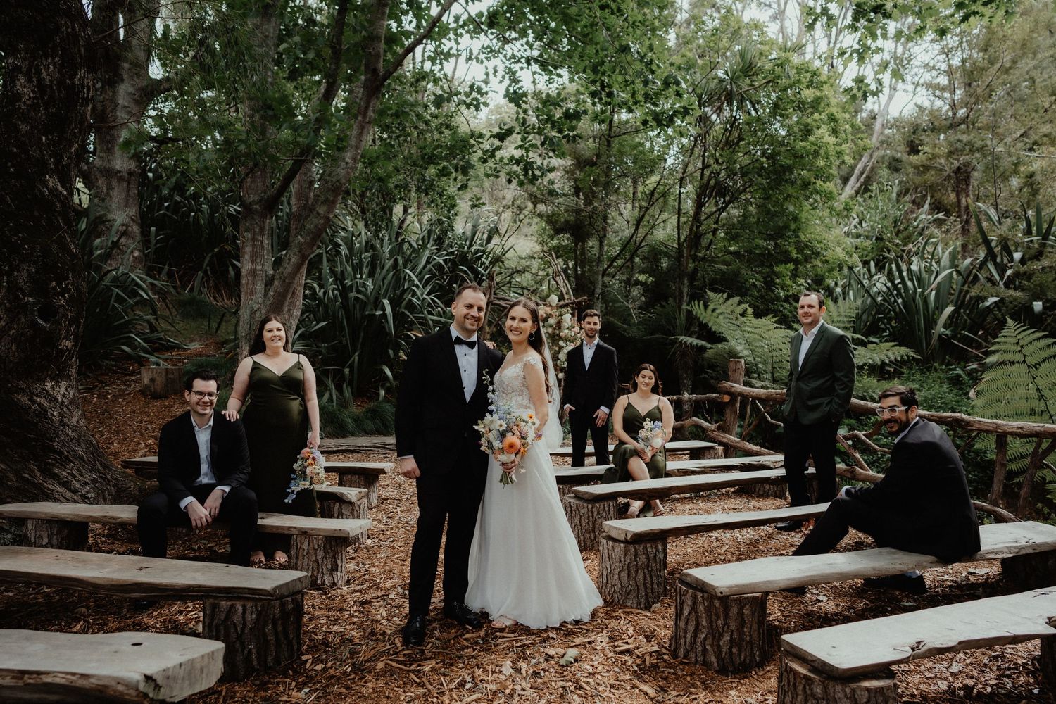 Wedding party in formal attire gathers on rustic wooden benches around the bride and groom in a lush forest garden ceremony.