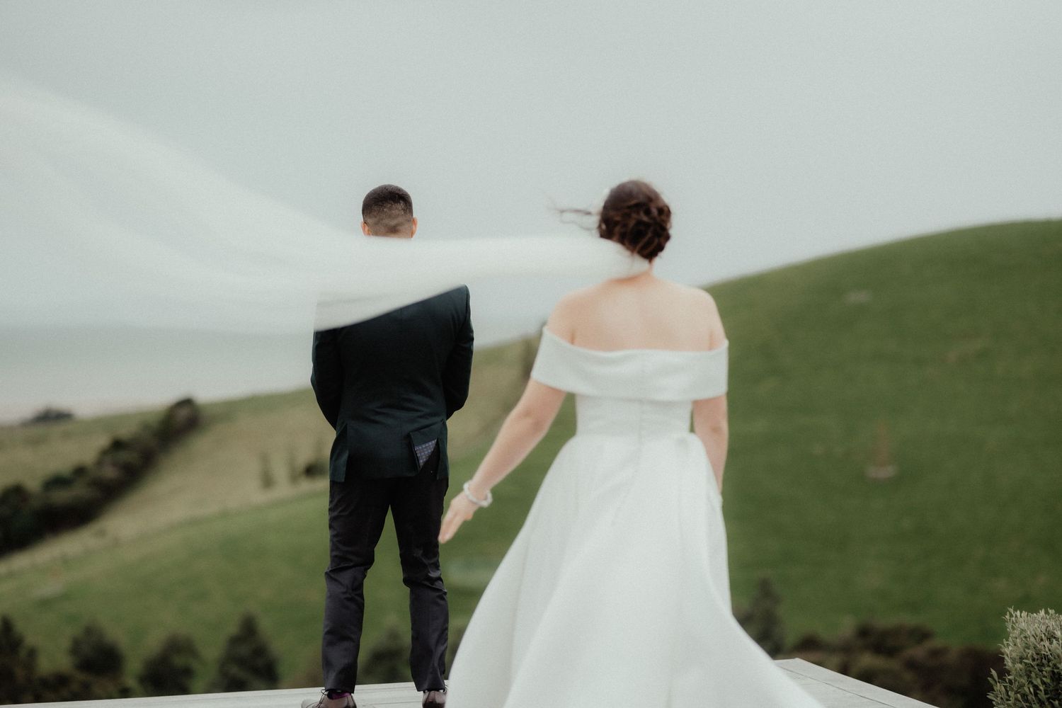 Bride in an off-shoulder white gown stands behind the groom as her veil blows across green rolling hills at a wedding.