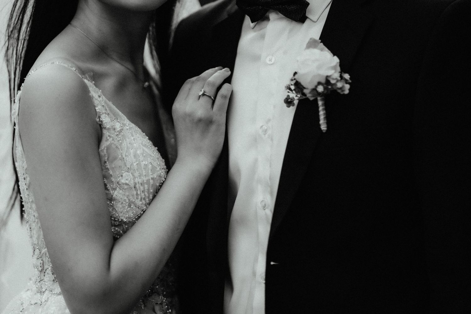 Black and white close-up of a bride's hand with a ring resting on a groom's chest, showing his boutonniere and bow tie.