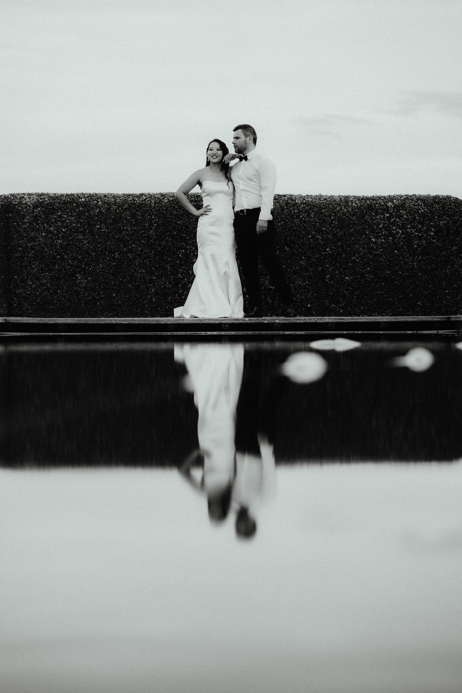 Black and white photo of a bride and groom embracing on a ledge before a tall hedge, reflected in still water.