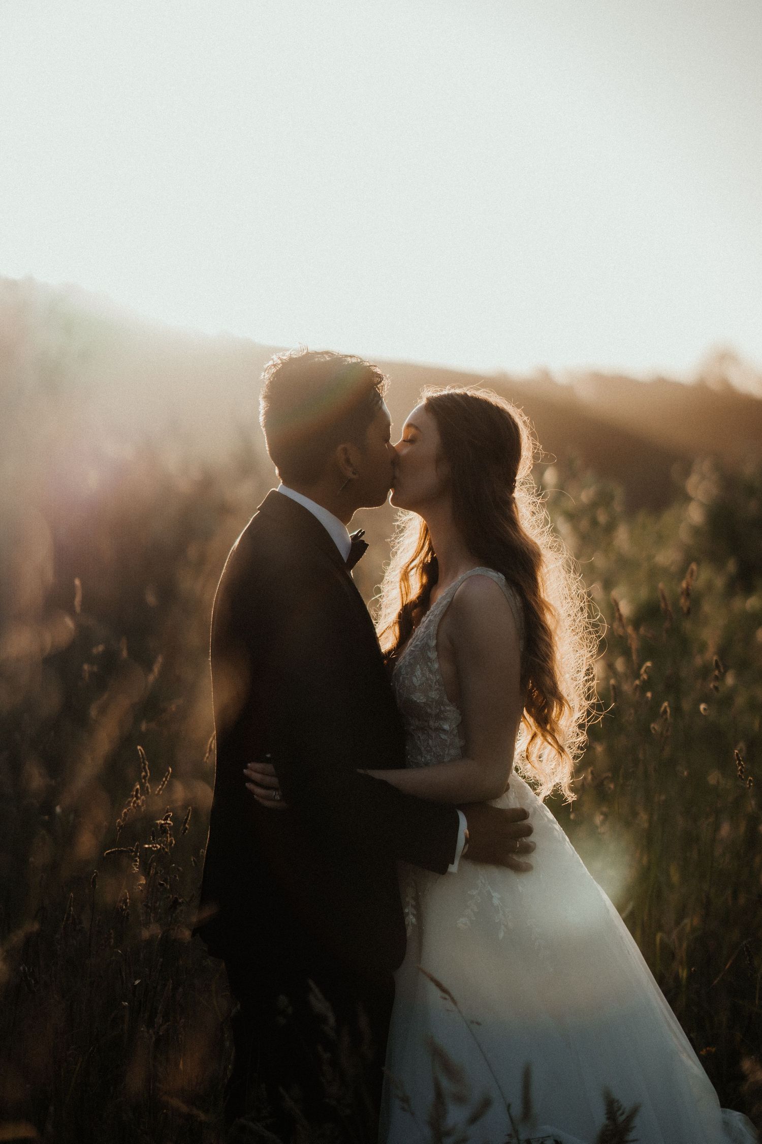A bride and groom share a romantic kiss in a golden field as warm sunset light glows behind them.