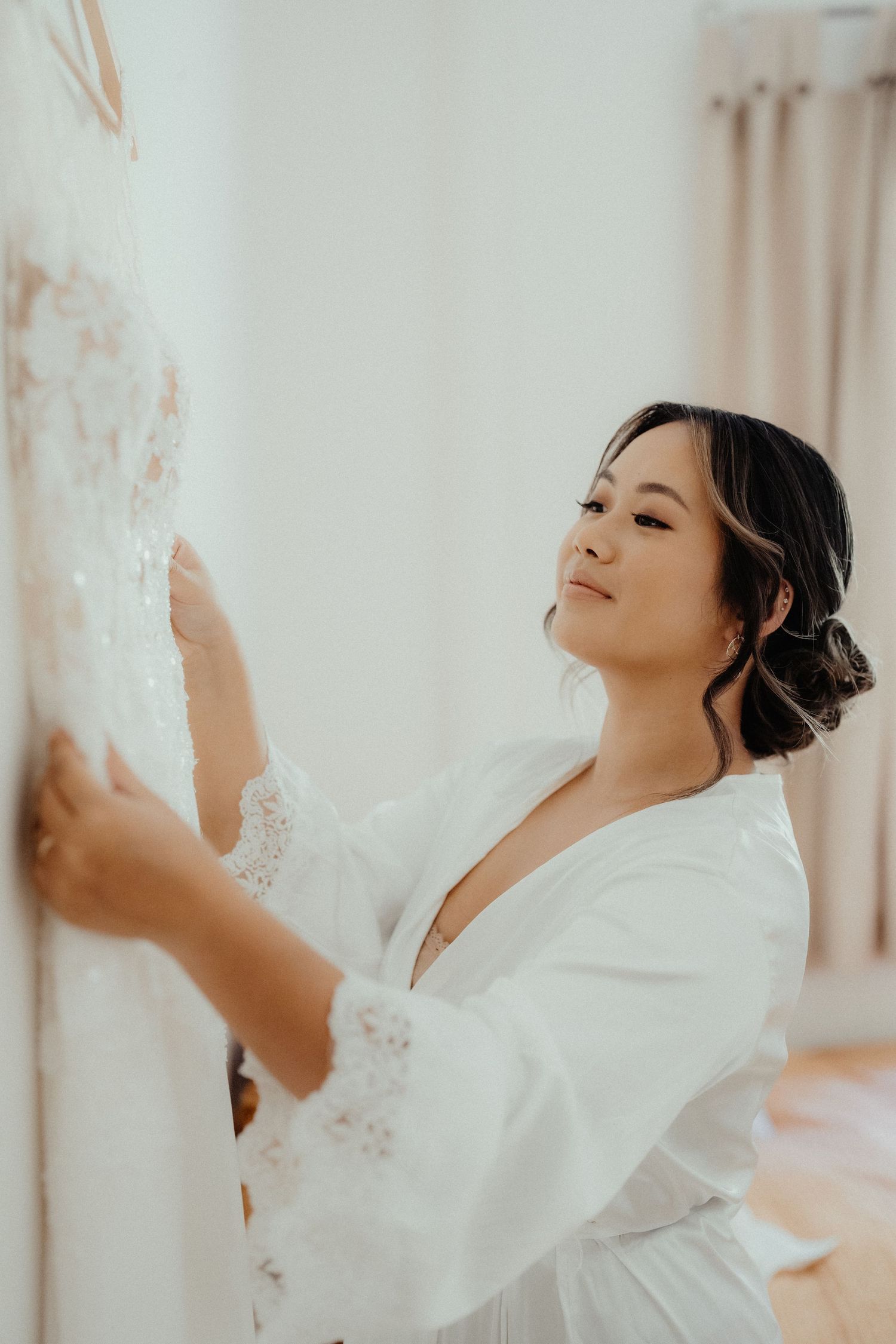 Bride in a white lace robe gently touches her beaded wedding gown hanging on the wall while getting ready.