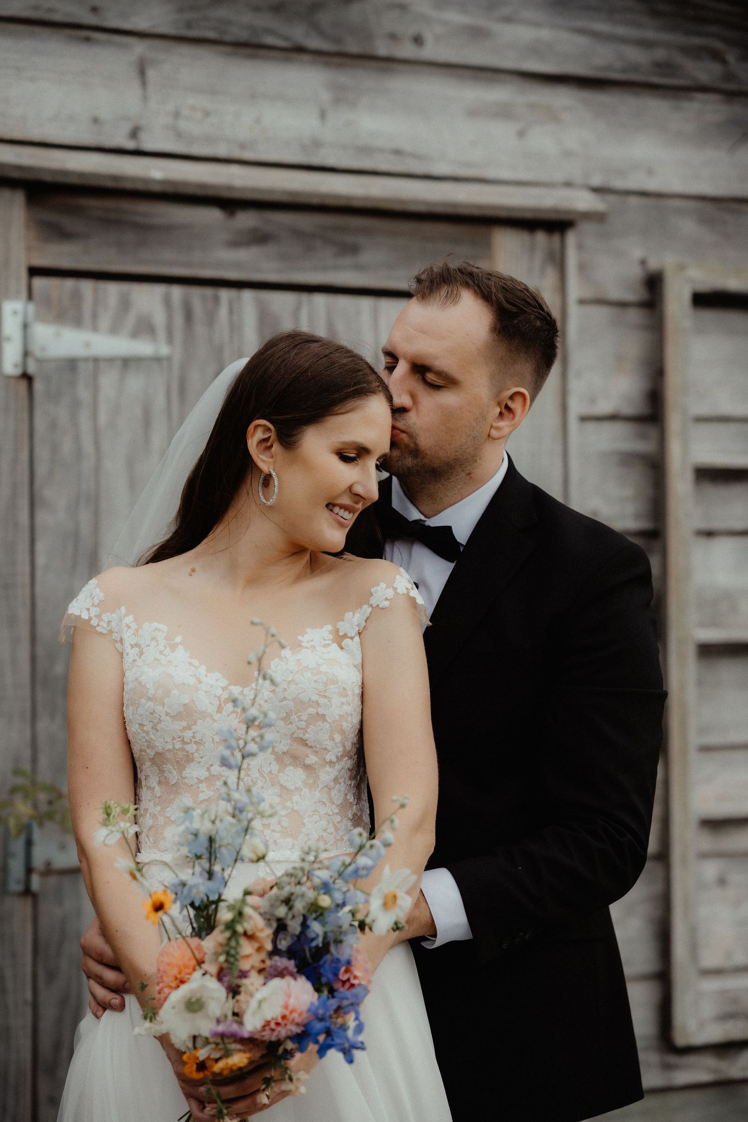 Groom kisses his smiling bride on the forehead as she holds a colorful wildflower bouquet before a rustic wooden barn door.