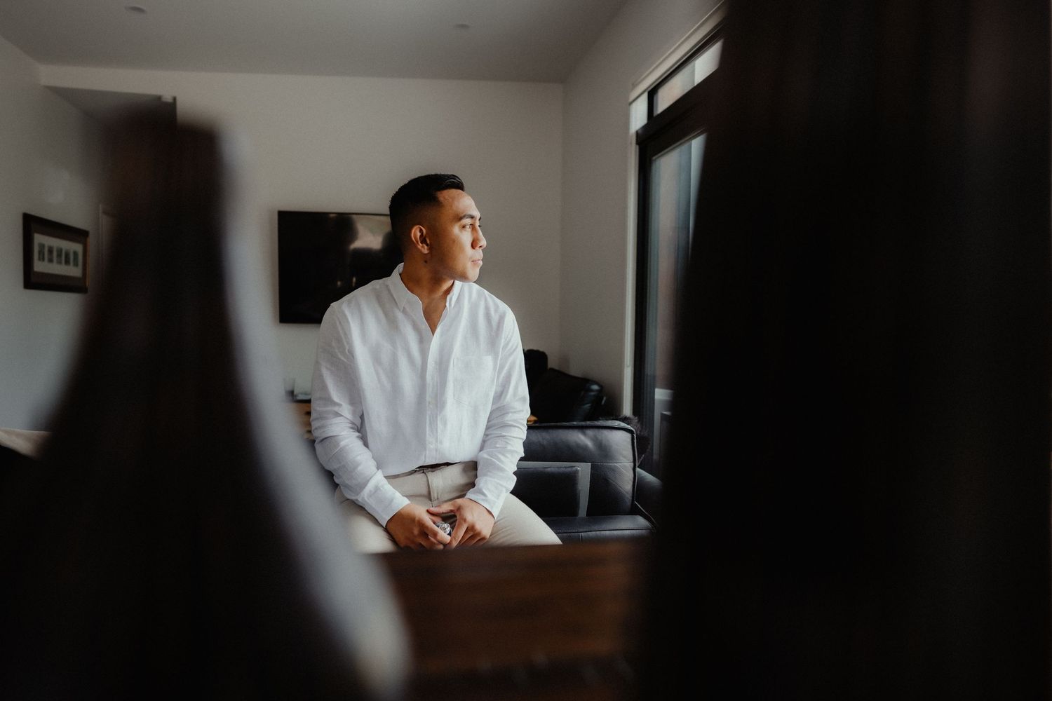 Groom in a white shirt sits thoughtfully by a window in a modern room, gazing out while preparing for his wedding day.