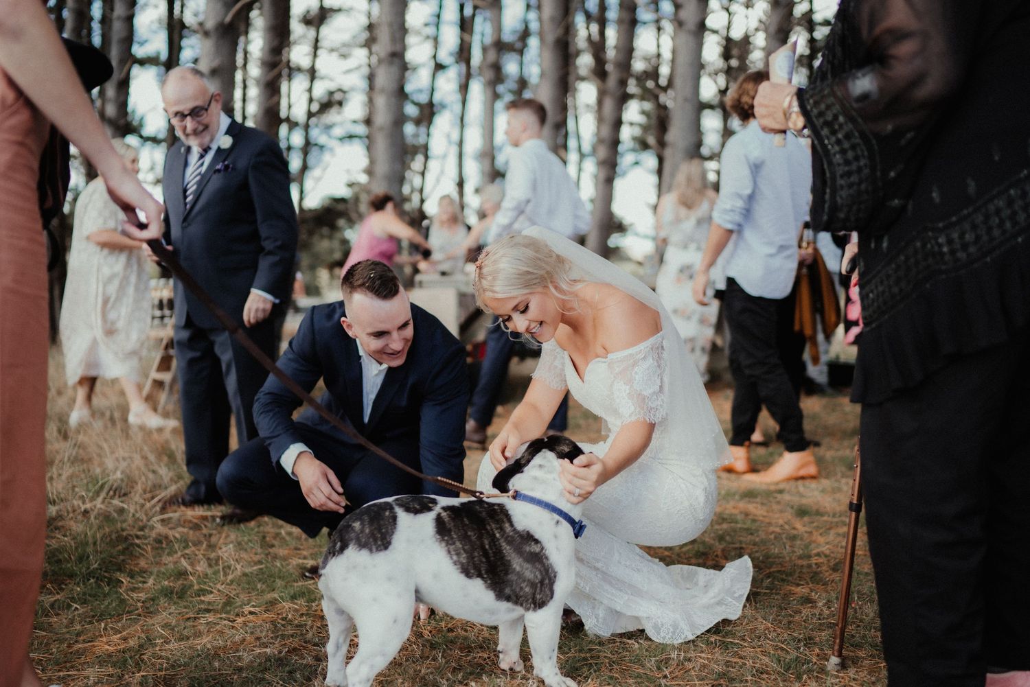 Bride in an off-the-shoulder gown holds a lush bouquet of white roses and eucalyptus leaves beneath trees at an outdoor wedding.