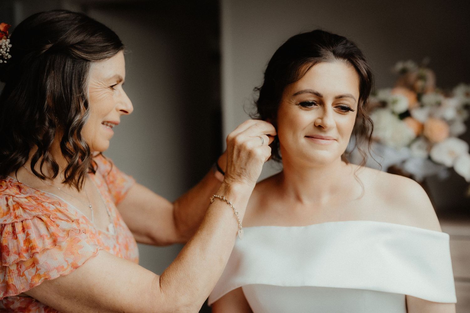 Mother helps the bride fasten an earring as they smile together, with the bride in a white off-the-shoulder wedding dress.