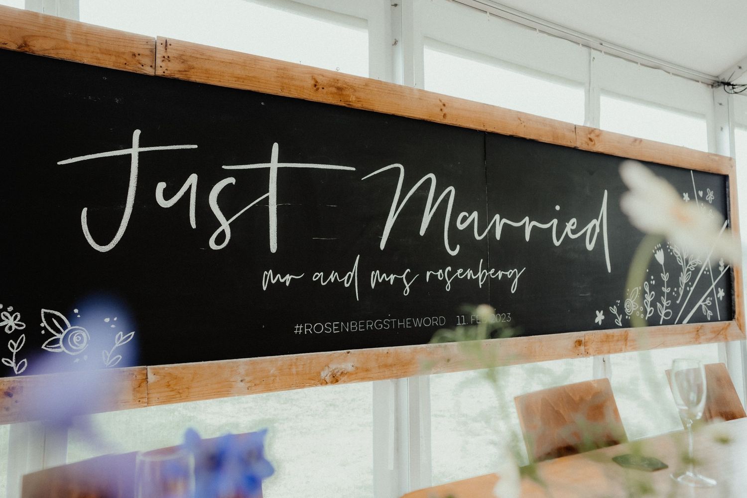 A chalkboard sign reading Just Married, Mr and Mrs Rosenberg, in a rustic wooden frame with soft flowers in the foreground.