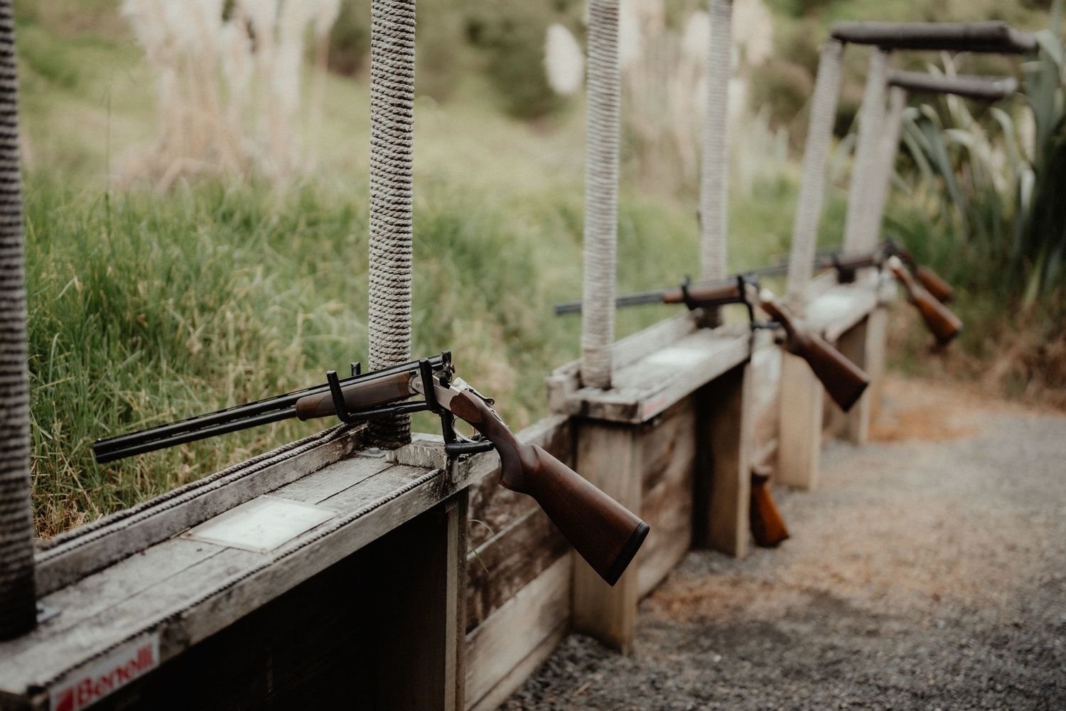 Vintage-style rifles mounted on a rustic wooden shooting stand beside a grassy hillside at an outdoor wedding venue.