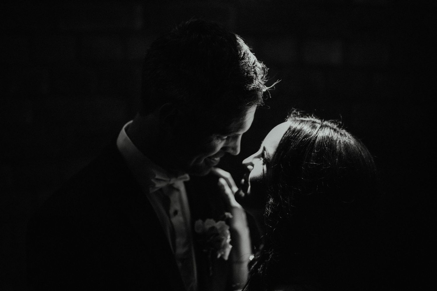 Moody black and white portrait of a couple about to kiss, their faces softly lit against a dark background.