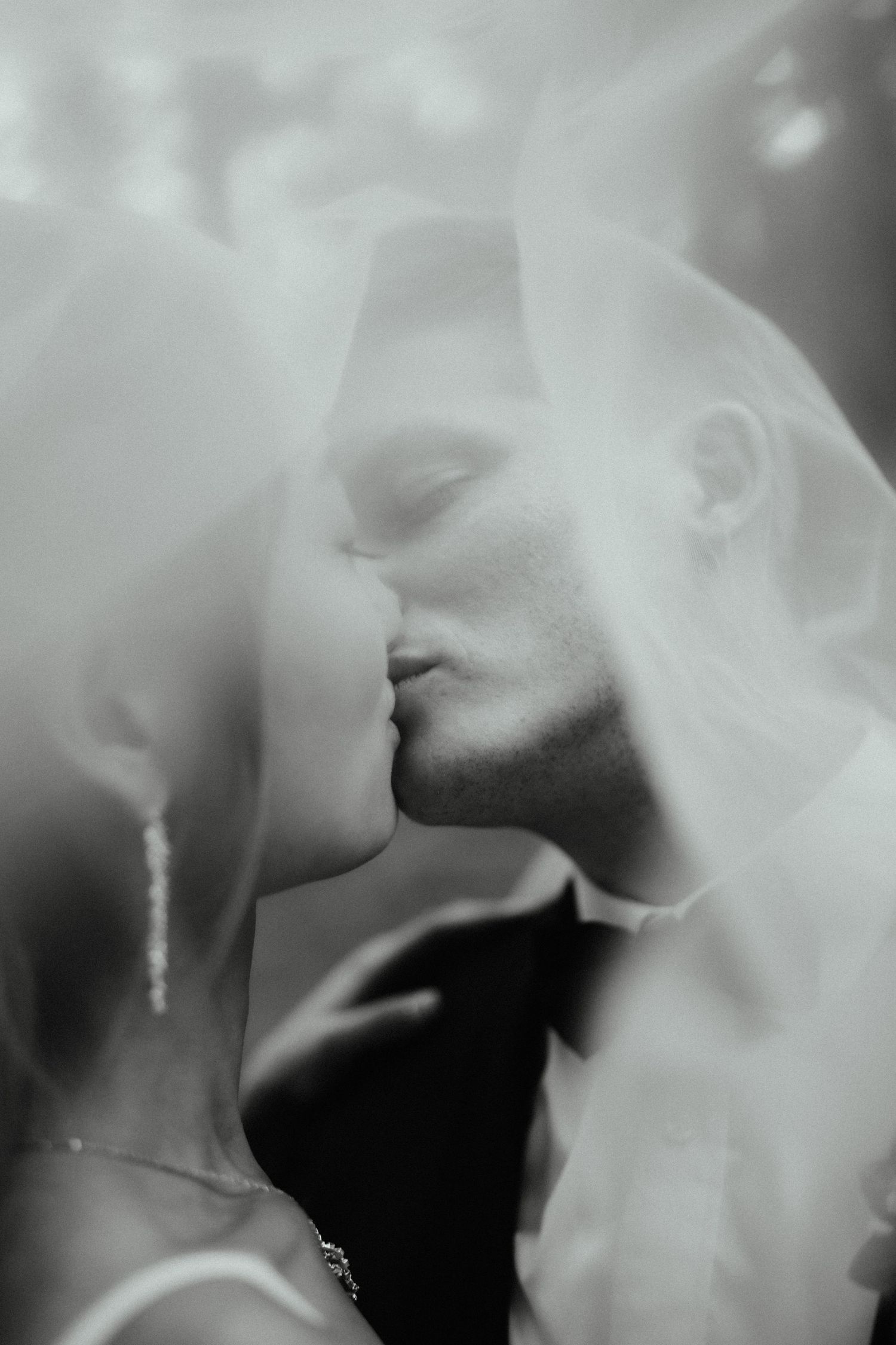 Close black and white wedding portrait of a bride and groom kissing softly beneath a sheer veil.