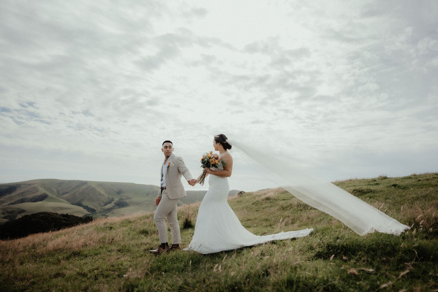 Bride in a white gown with a flowing veil holds hands with the groom as they walk across a grassy hilltop under a cloudy sky.