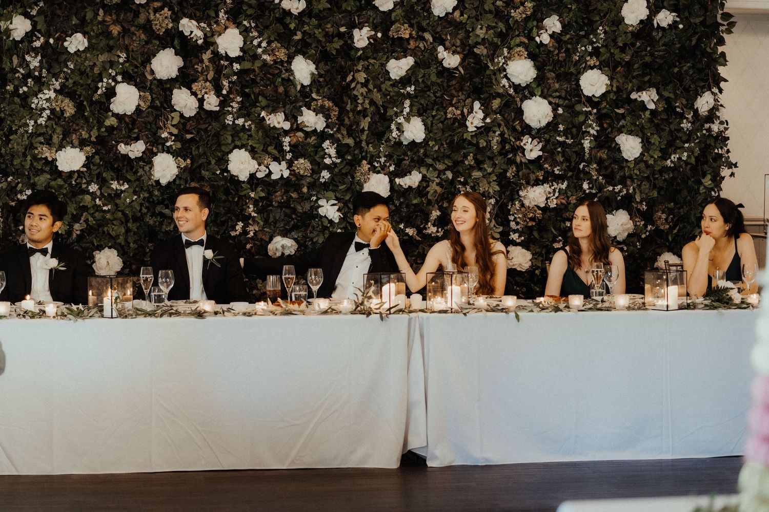Wedding party seated at a long head table in front of a white floral wall, with glasses and candles set along the white linen.