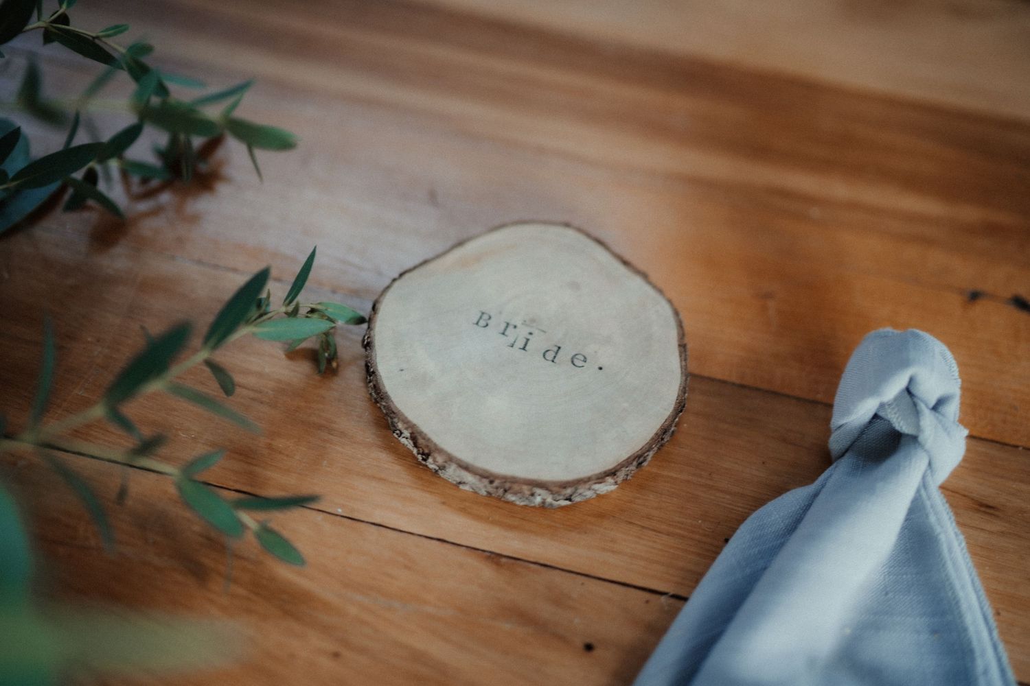 Round wooden slice engraved with the word bride sits on a timber table beside eucalyptus leaves and a blue napkin.