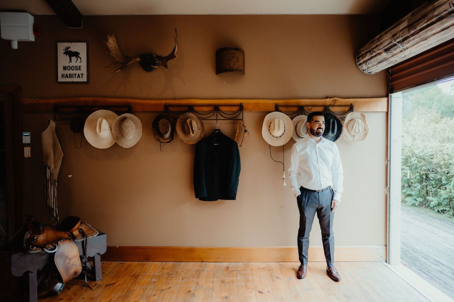 Groom in a white shirt and grey trousers stands in a rustic room beside a row of hats and a dark jacket hanging on the wall.