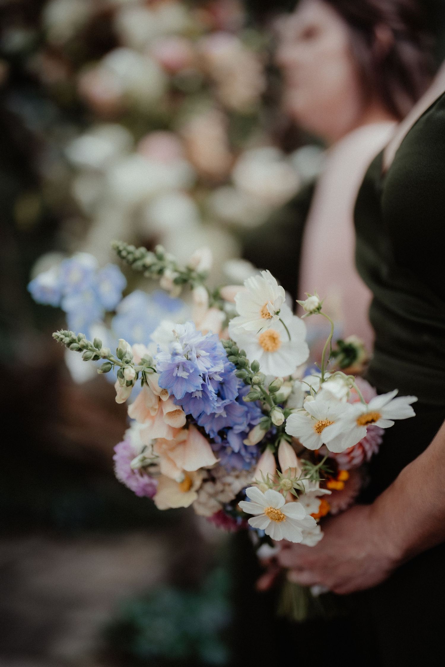 Close-up of a wildflower wedding bouquet with blue hydrangeas, white cosmos and pink blooms held by a bridesmaid in a dark green dress.
