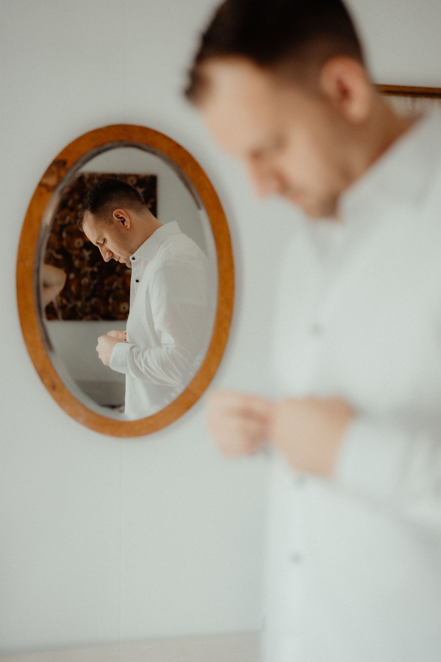 Groom in a white shirt buttons his cuff while getting ready, his reflection visible in an oval wood-framed mirror.