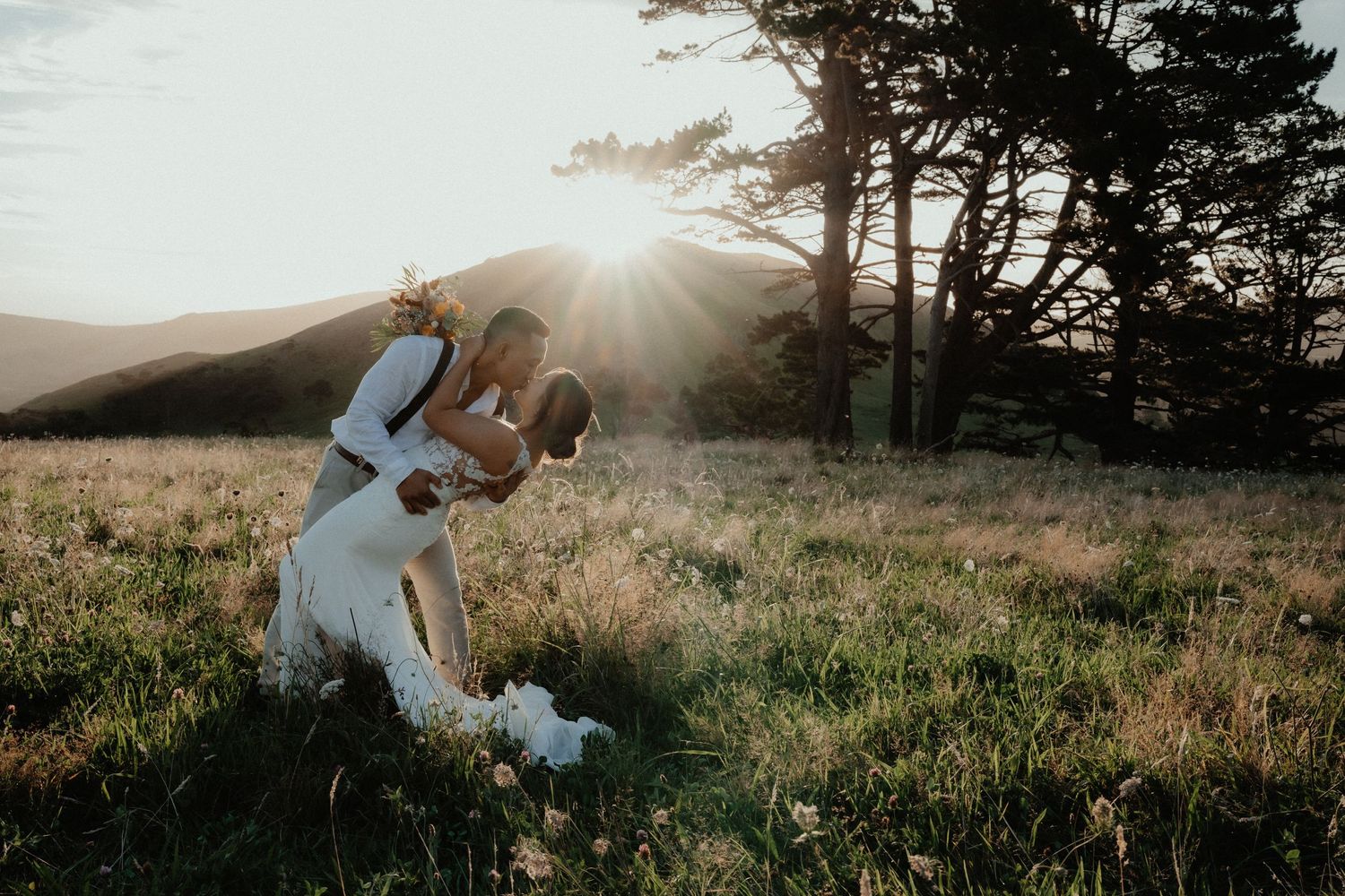Groom dipping bride in a romantic kiss in a grassy field at sunset, with hills and trees in the golden background.