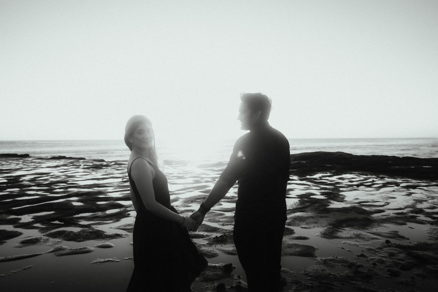 Silhouetted couple holds hands facing the glowing sun on a rocky shoreline in a black and white beach engagement photo.