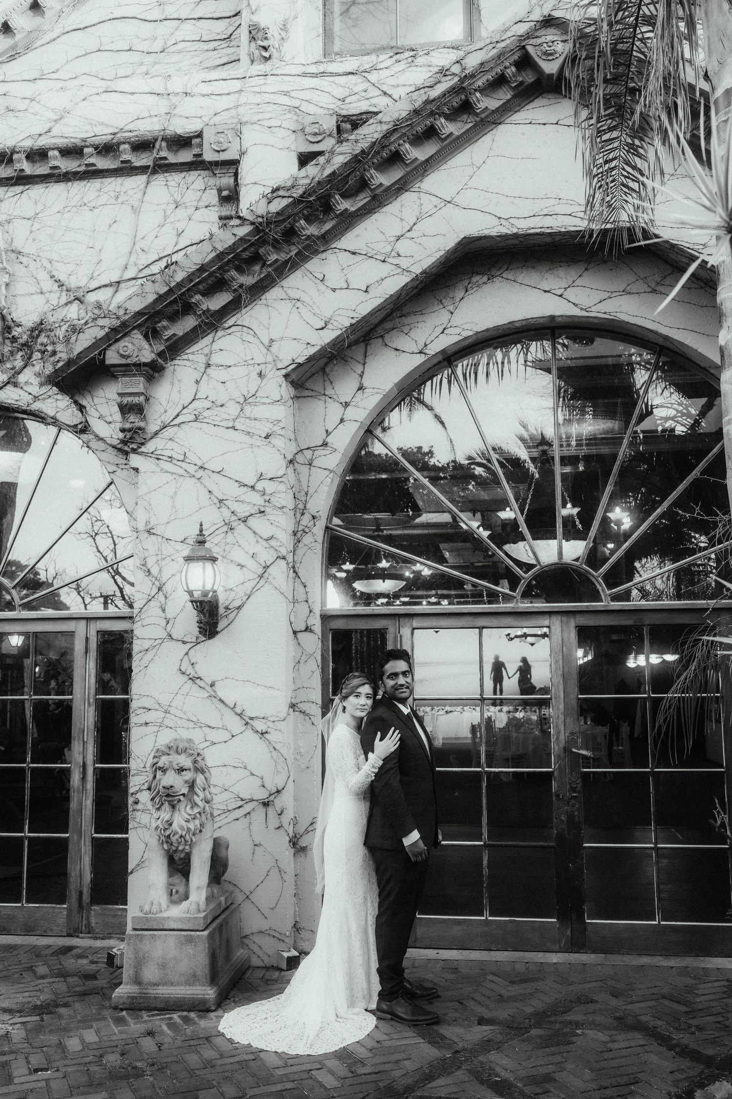 Black and white portrait of a bride and groom embracing in front of a vine-covered building with arched windows and a lion statue.