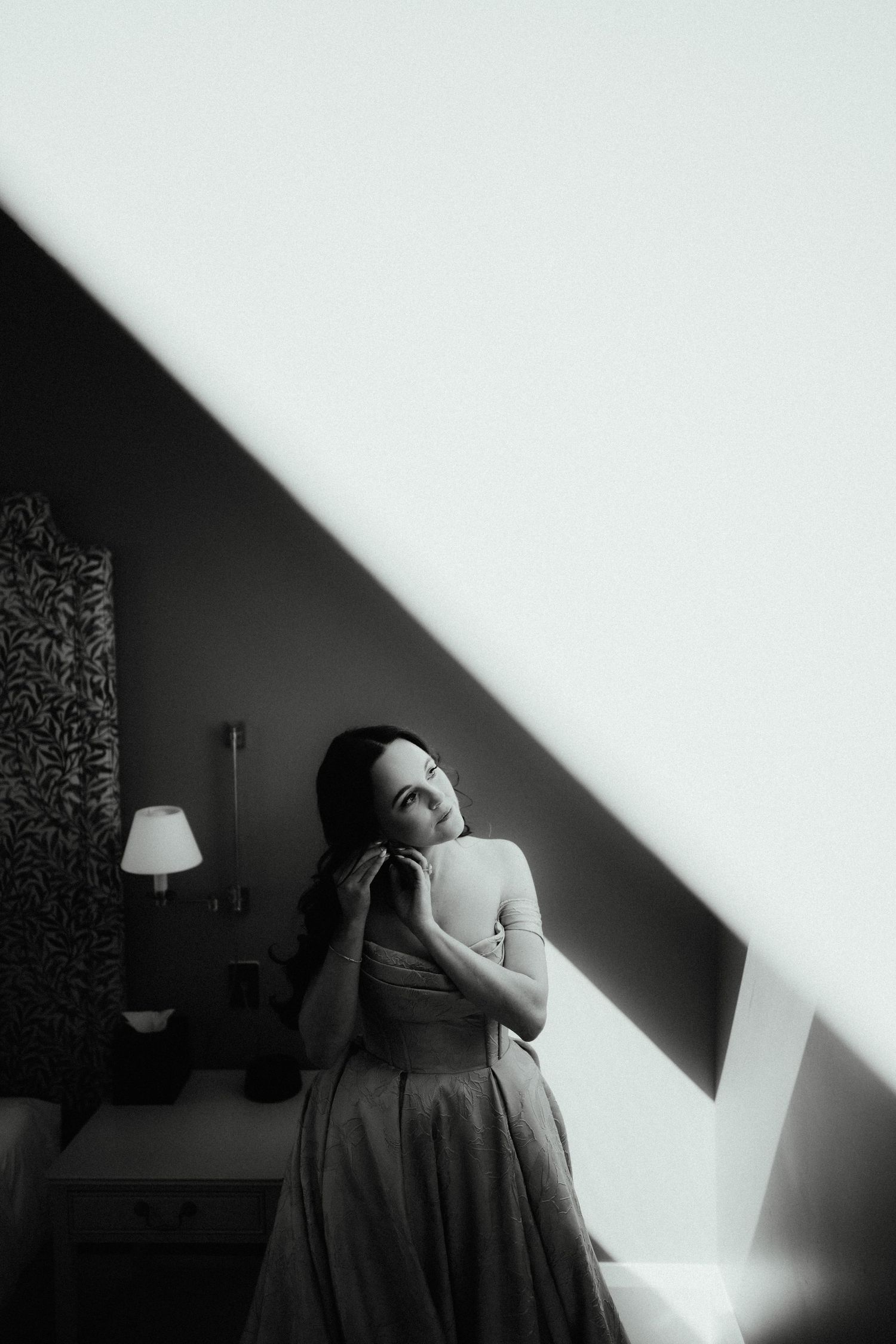 Black and white portrait of a bride in a strapless gown touching her earring beside a window with dramatic light and shadow.