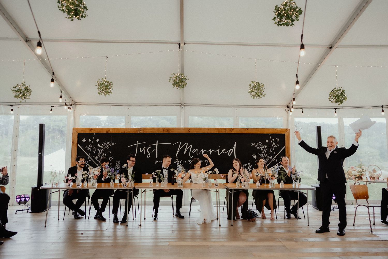 A man cheers beside the head table in a marquee reception, with a Just Married chalkboard sign behind the bridal party.