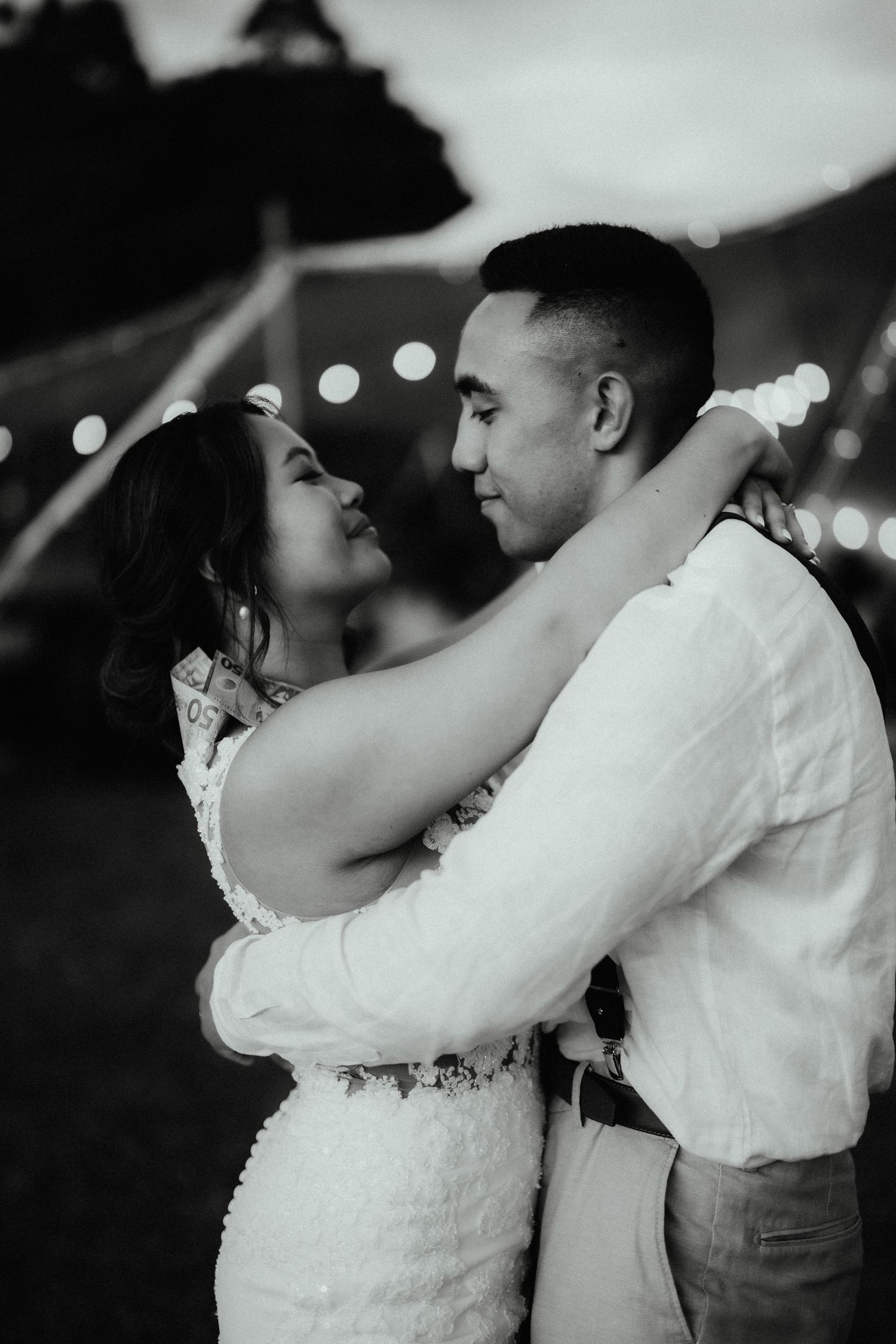 A black and white photo of a bride and groom embracing and gazing at each other under string lights at their evening reception.