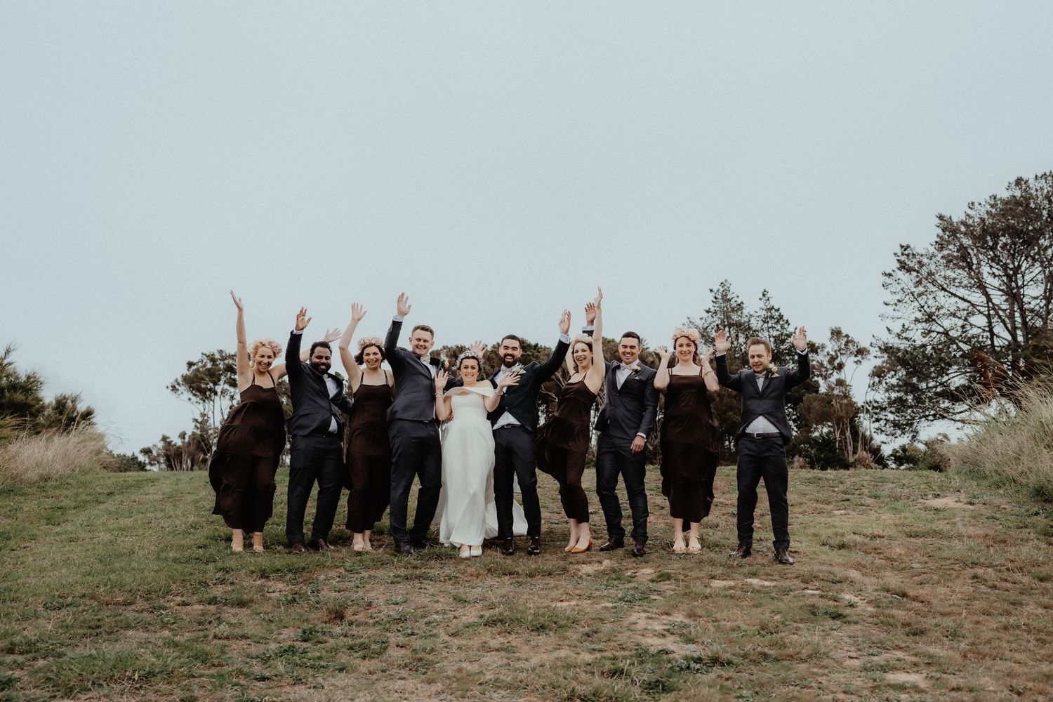 Joyful bridal party raises their arms in celebration while the bride in a white gown stands center on a grassy hillside.