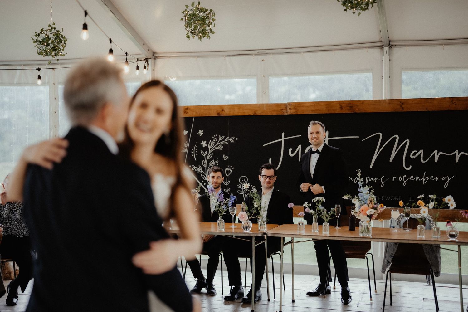 Bride dances with an older man during a wedding reception while the groom and guests watch from tables in a decorated marquee.