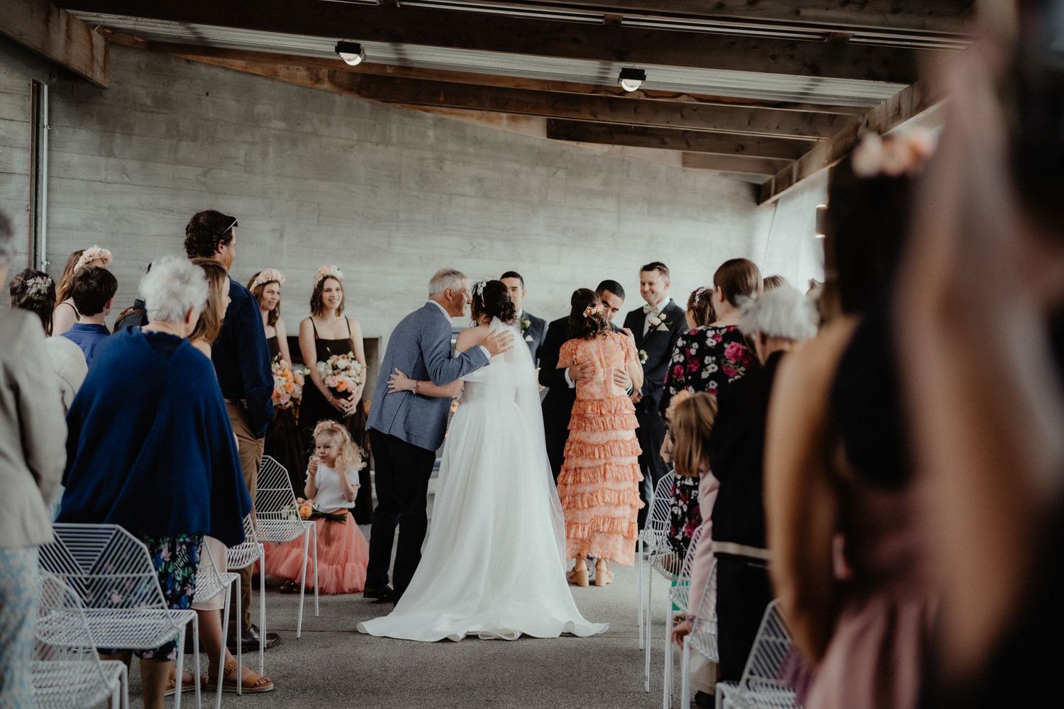 Bride in a white gown walks down the aisle with an older man as seated wedding guests in colorful dresses look on.