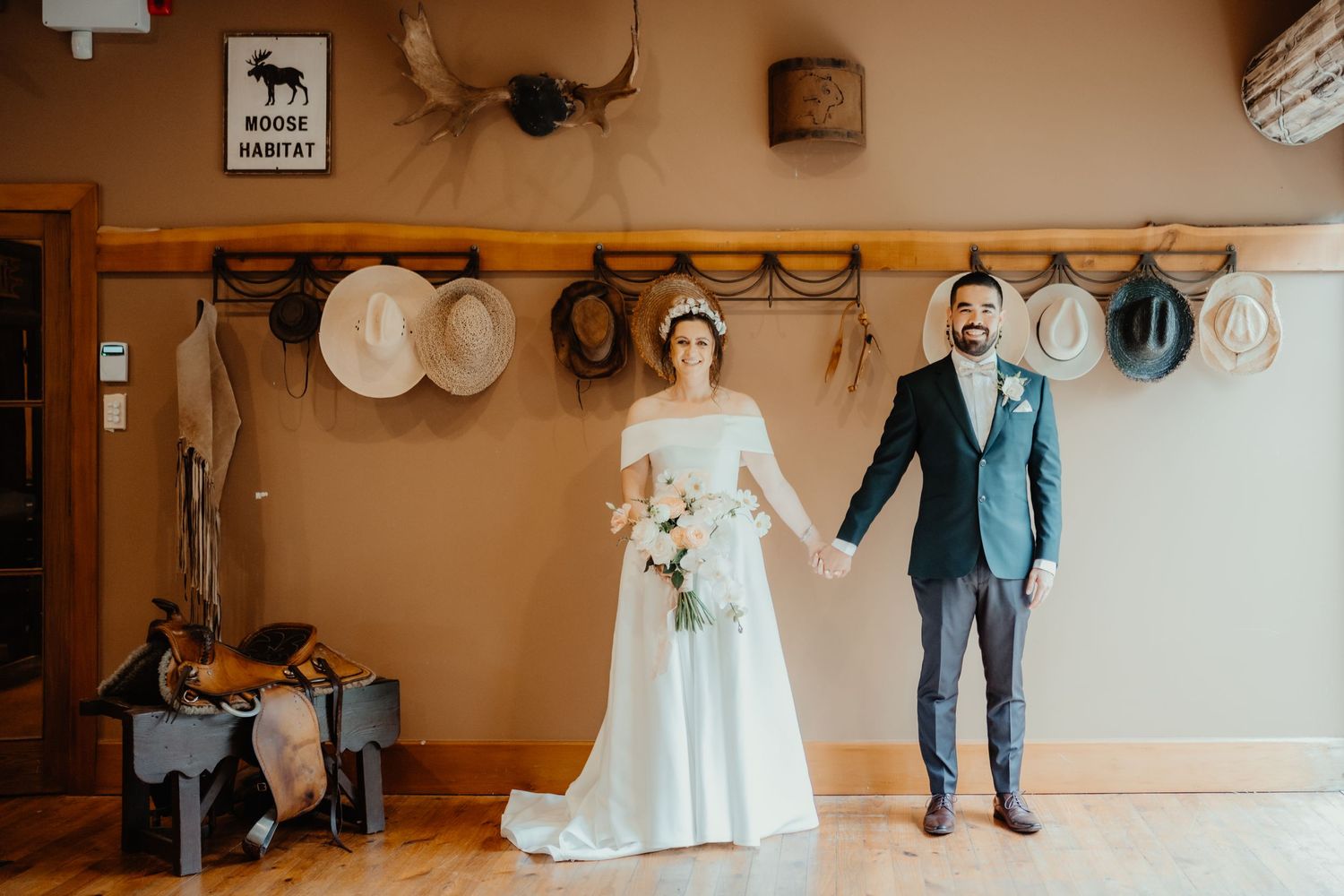 Bride and groom hold hands in a rustic room with hats hanging on a wooden rail, the bride holding a bouquet in a white gown.