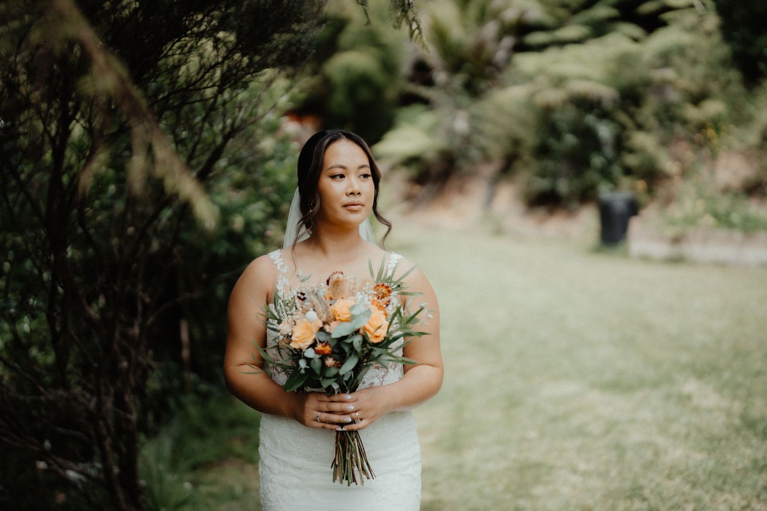 Bride in a white lace dress holds an orange and peach bouquet while standing outdoors among lush green foliage at her wedding.