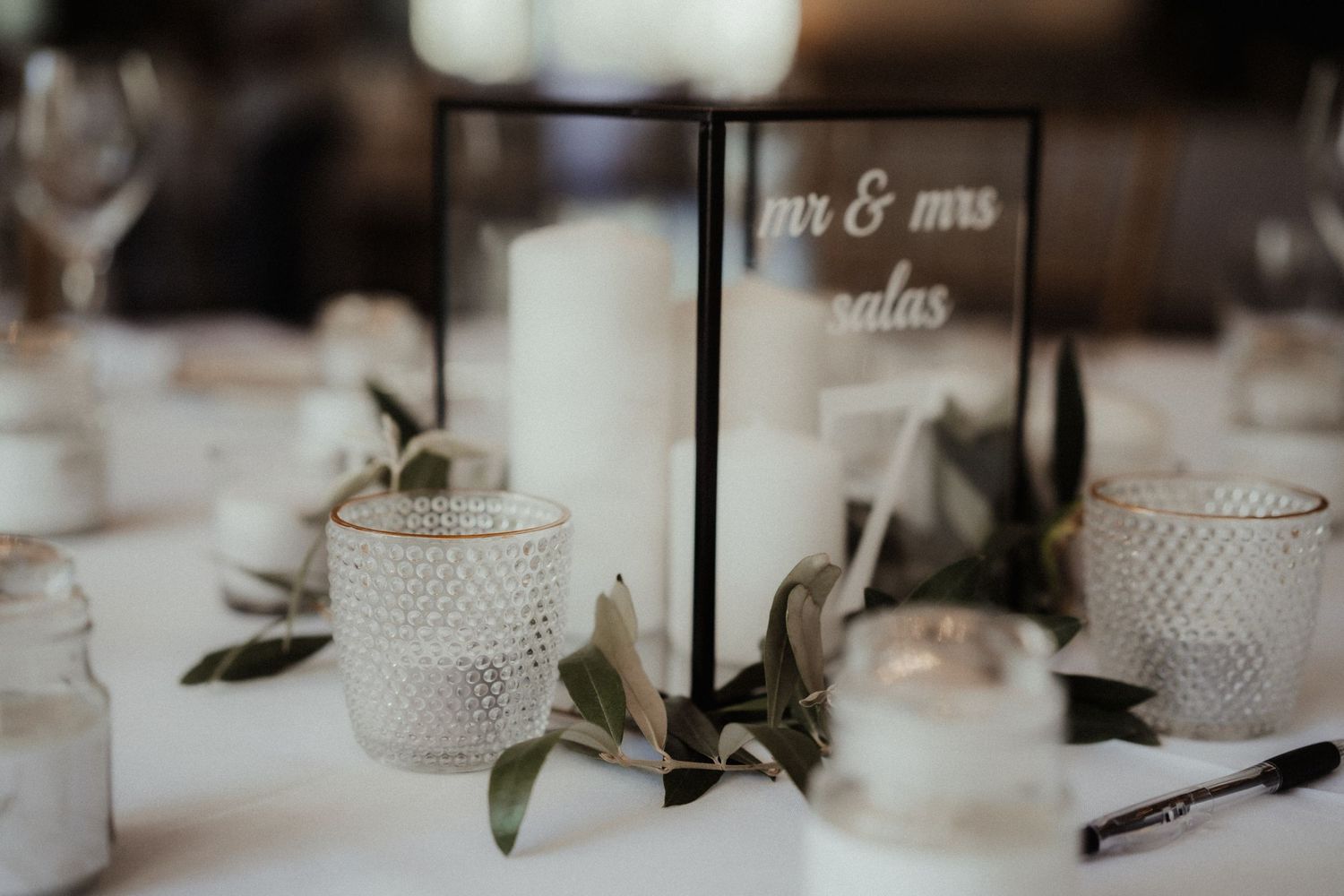 A framed Mr and Mrs table sign sits among glass candle holders and olive branch greenery on a white wedding reception table.