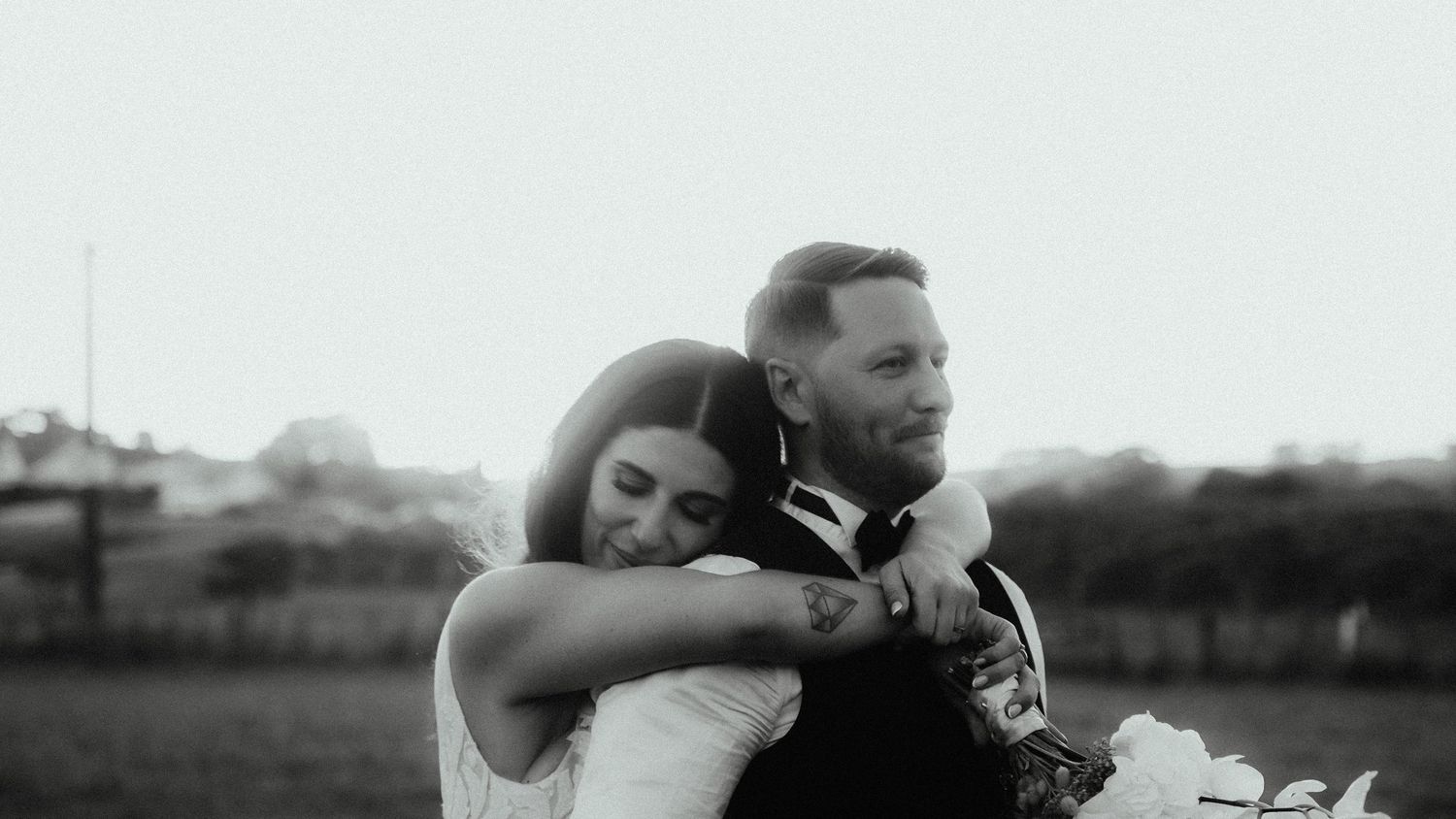 Black and white photo of a smiling bride hugging her tattooed groom from behind while holding a bouquet outdoors.
