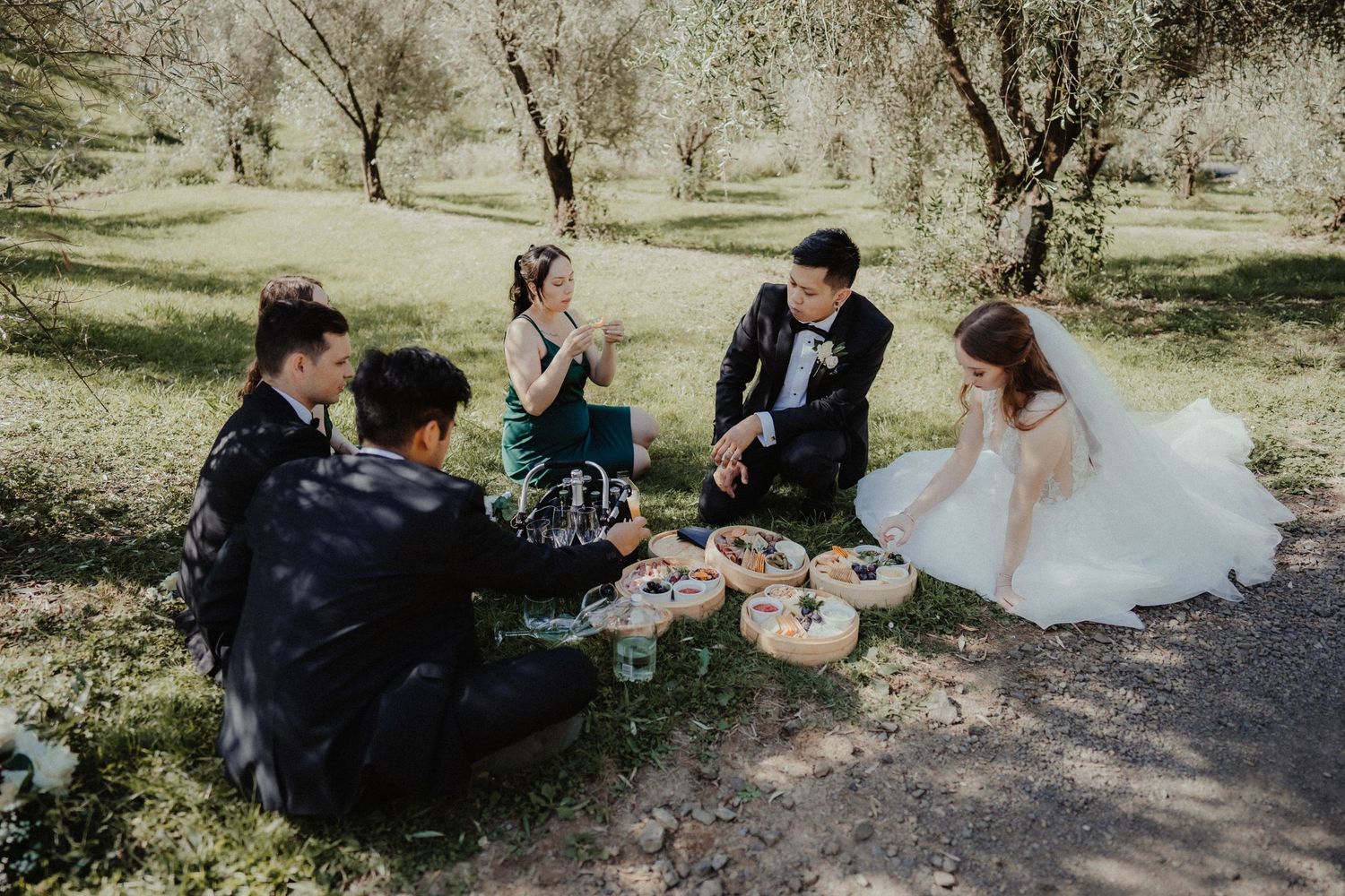 Wedding guests sit on the grass in an olive grove sharing a picnic while the bride in a white gown sits nearby.