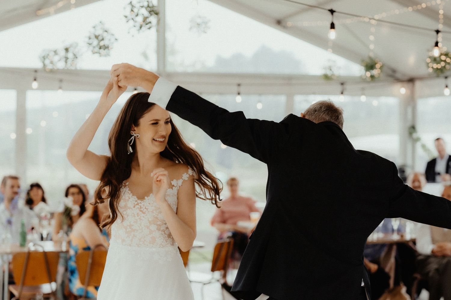 A joyful bride in a lace dress twirls under her partner's raised arm during a dance at a wedding reception inside a marquee.