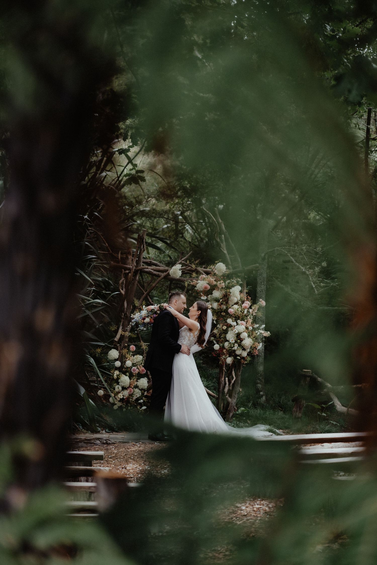 Couple shares an intimate moment before a rustic wedding arch decorated with white flowers, seen through soft green forest foliage.