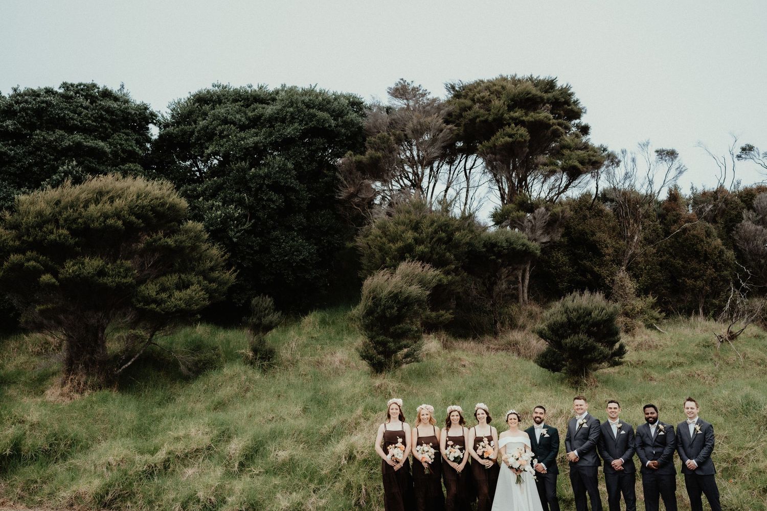 Wedding party of bridesmaids in brown dresses and groomsmen in gray suits poses with the bride on a grassy hillside.