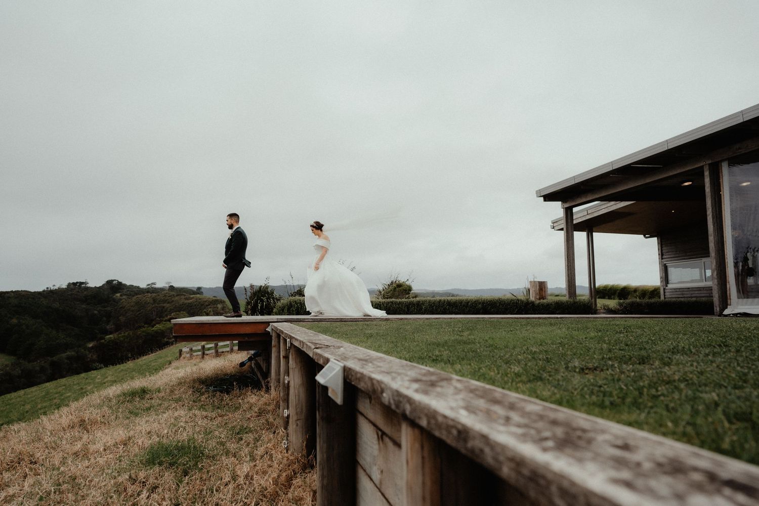 Bride in a flowing white gown and groom in a dark suit walk across a grassy hilltop on a moody overcast day.