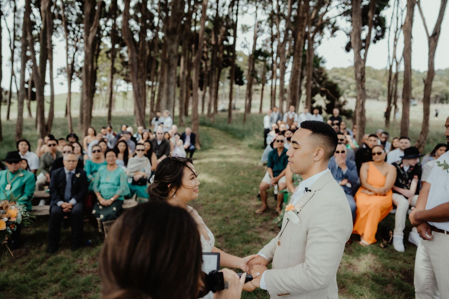 Wedding guests watch the couple exchange vows during an outdoor ceremony among tall trees on a grassy lawn.