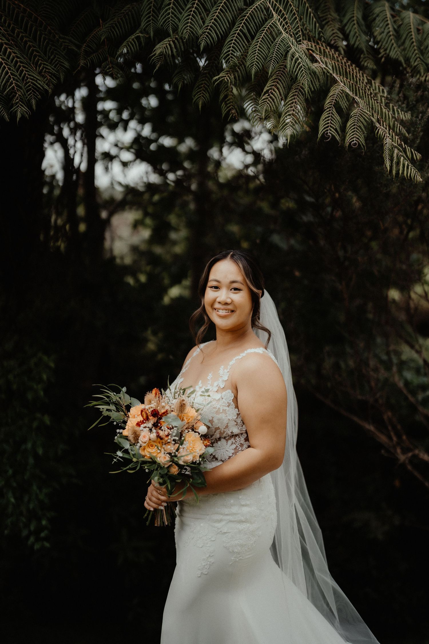 Smiling bride in a lace wedding gown and long veil holds an orange bouquet beneath fern fronds.