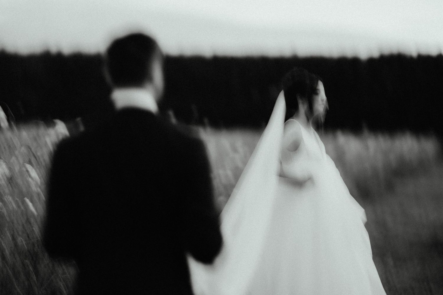 Blurred black and white wedding photo of a groom in a suit and a bride in a veil walking through a field.