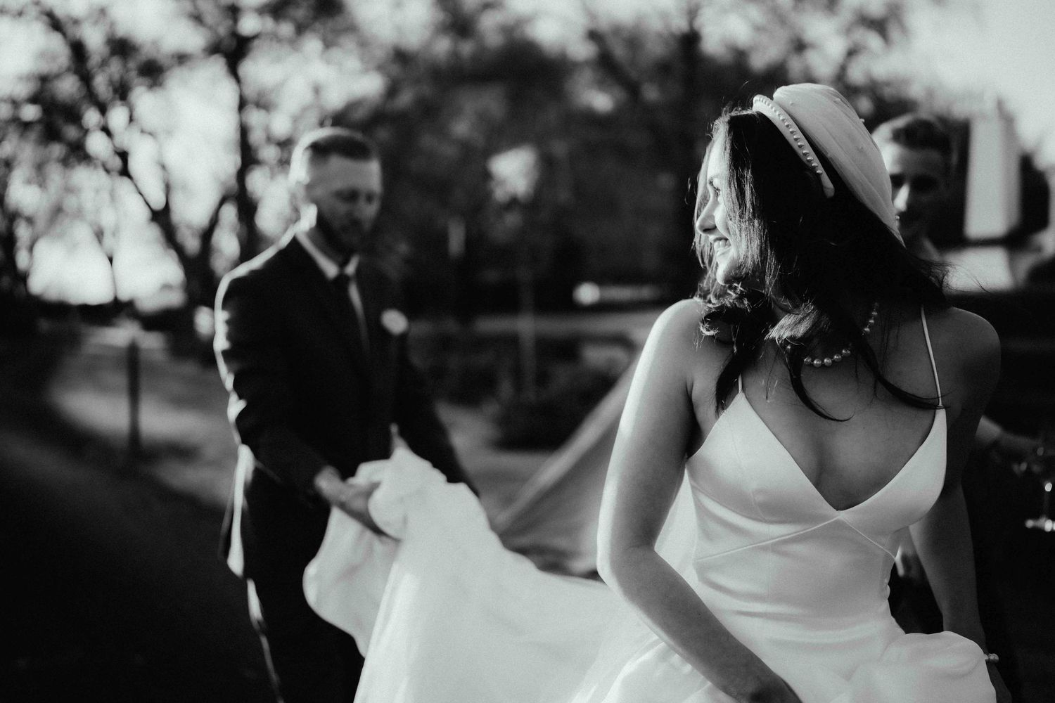 Black and white wedding photo of a bride in a V-neck gown and hat with a groom in a suit holding her flowing skirt outdoors.